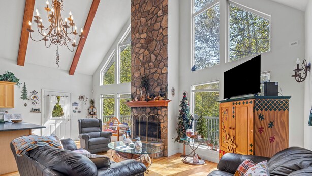 A spacious living room with a stone fireplace, leather sofas, a chandelier, and large windows offering a view of trees. - Cabin on the River - In Town on the Chattahoochee River (Helen)