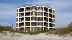 A multi-story building with a curved facade, balconies, and a sandy beach in the foreground.