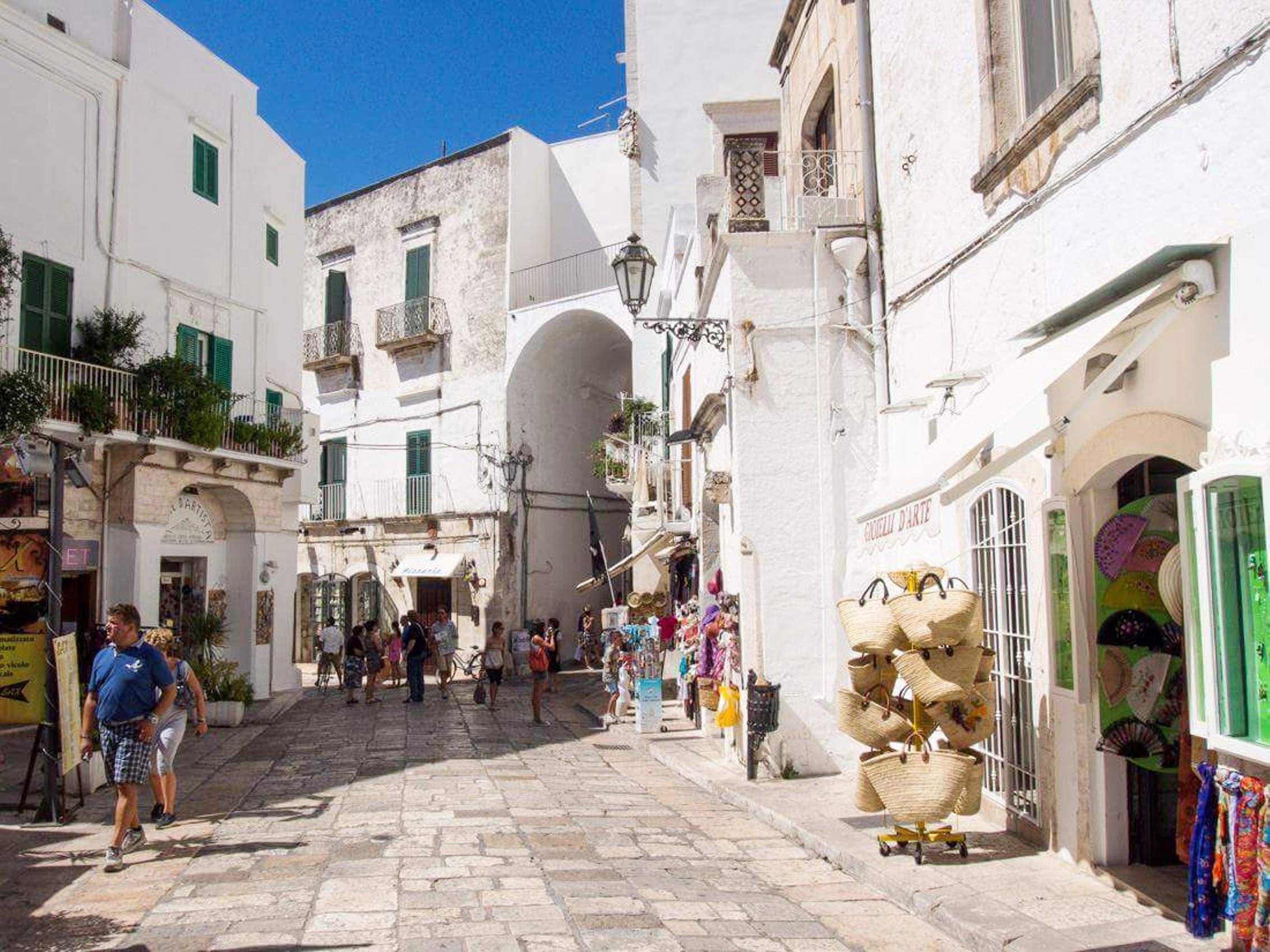 Una calle estrecha con edificios blancos, un arco central y una tienda que vende cestas.