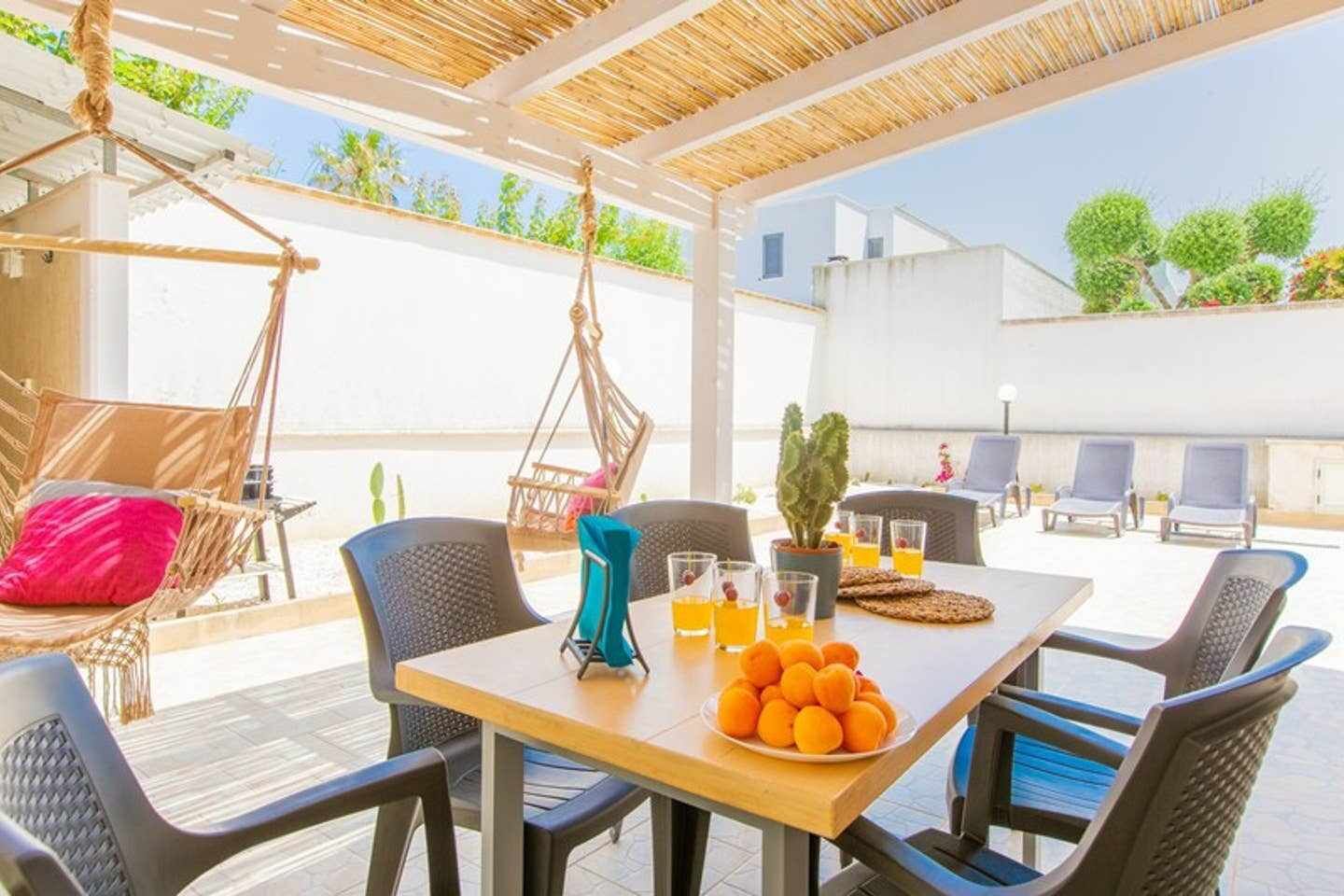 A covered outdoor seating area with a table set for two, surrounded by chairs, a hanging chair, and a cactus plant.
