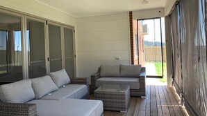 A sunlit patio with a grey sofa, a wicker ottoman, and a sliding glass door leading to a backyard.