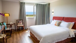 A hotel room with a large bed, a desk with a chair, a lamp, and a view of the sea through the window.