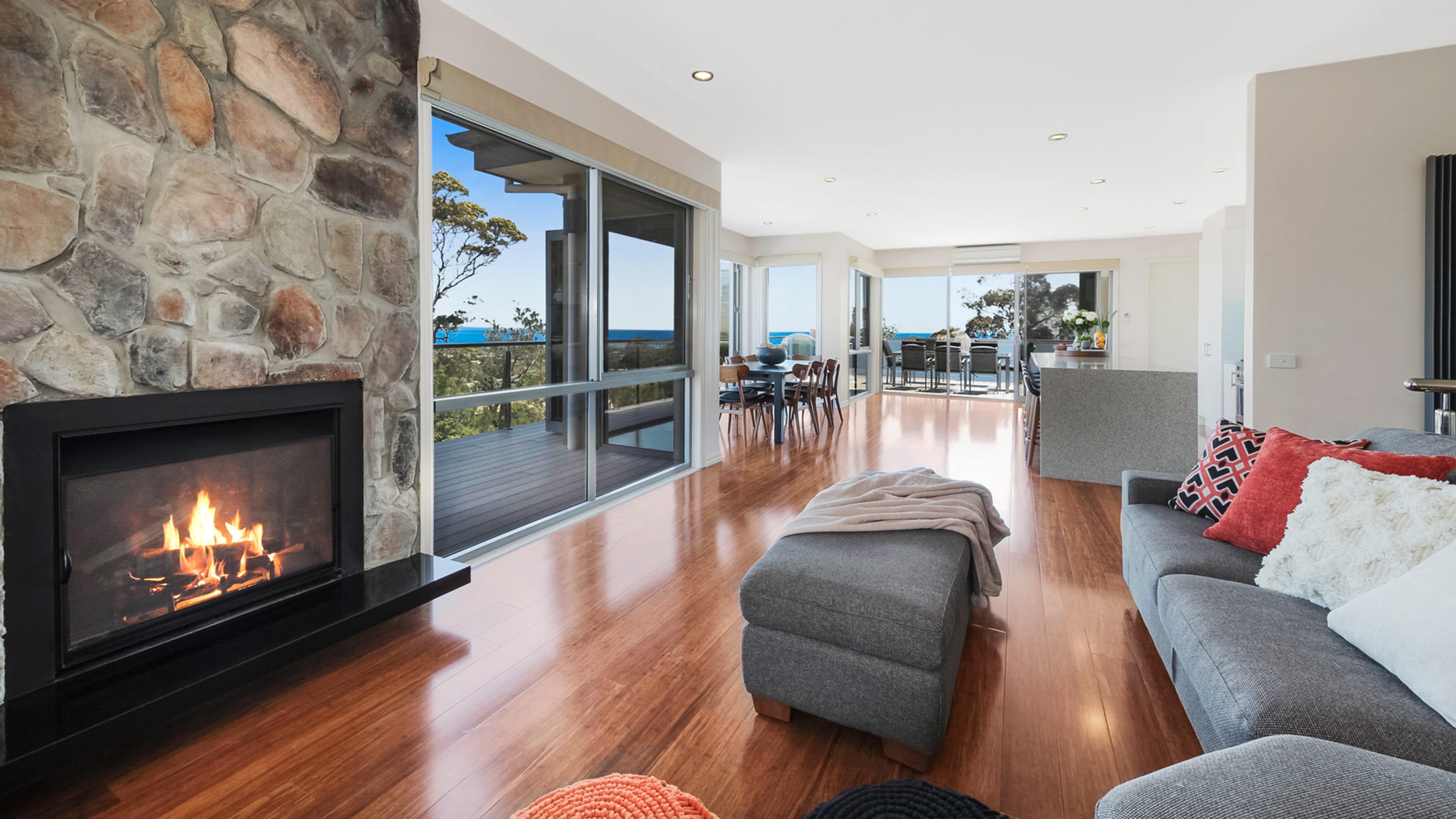 A modern living room with a stone fireplace, a grey sofa, and a view of the ocean.
