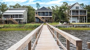 Un paseo marítimo de madera que conduce a una propiedad frente al agua con varias edificaciones, incluyendo una casa grande con balcón y una estructura más pequeña.