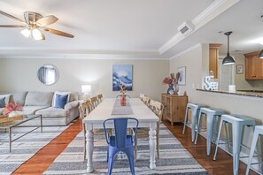 A spacious living room with a large dining table, blue chairs, a sofa, and a ceiling fan.