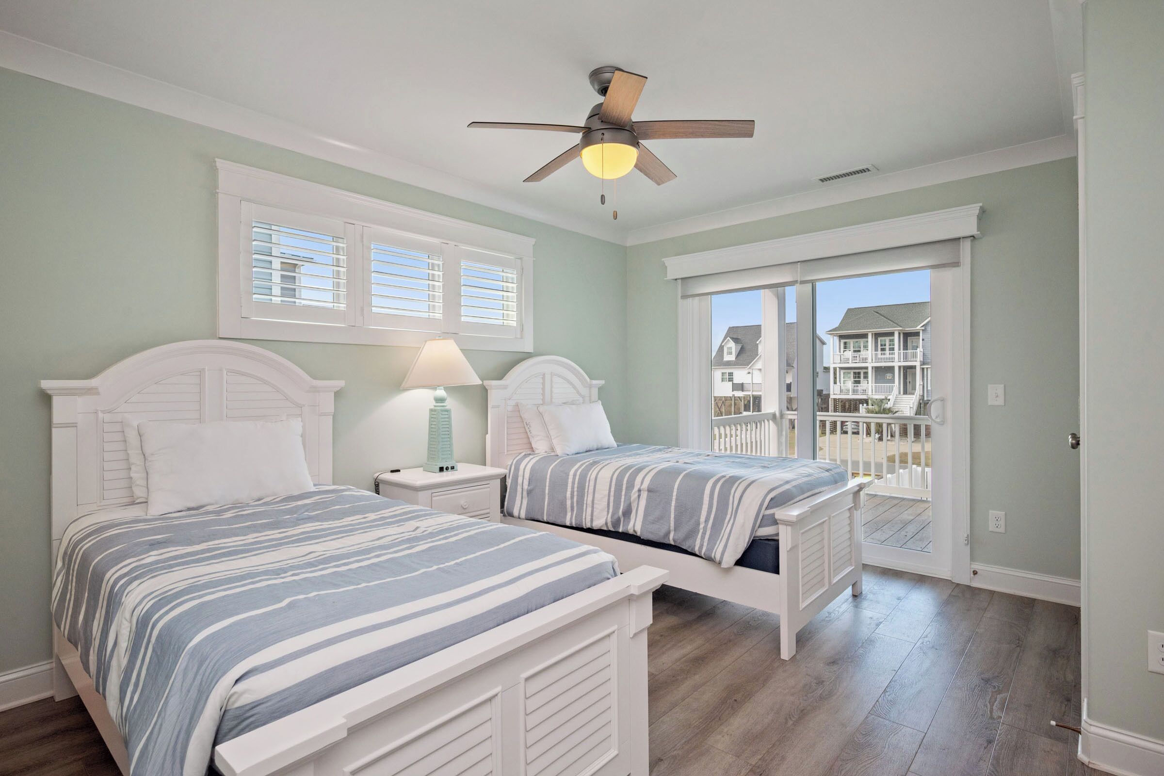 A bedroom with two beds, a ceiling fan, and a view of a house through a sliding glass door.