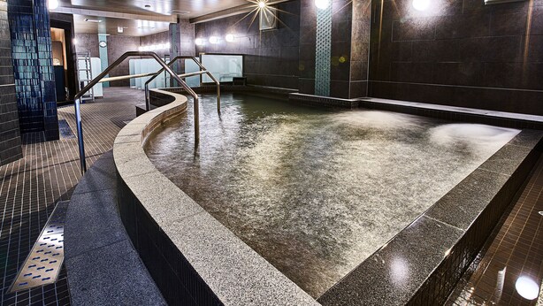 A modern indoor pool with a curved design, a central water feature, and a metal staircase leading to a higher level. -  Anshin oyado Woman & Man Nagoya (Nagoya)
