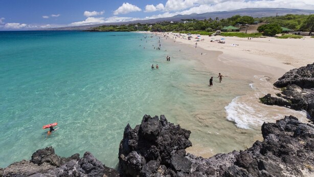 Ein Strand mit klarem türkisblauem Wasser, felsiger Küste und einem Sandstrand mit Menschen und Sonnenschirmen.