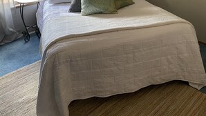 Ein ordentlich bezogenes Bett mit weißer Daunendecke und mehreren Kissen, ein Gemälde eines Piers an der Wand und ein Fenster mit durchsichtigen Vorhängen.