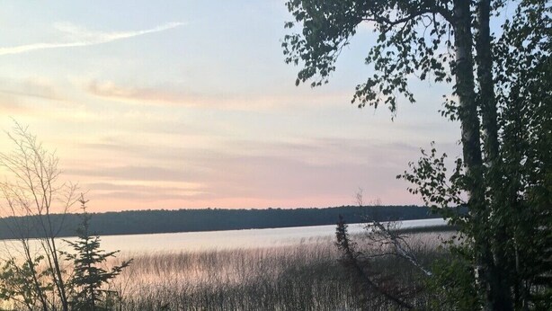 Ein Lagerfeuer mit drei Adirondack-Stühlen darum, gelegen auf einer Wiese mit einem See und Bäumen im Hintergrund.