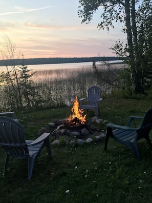 Een kampvuur met drie Adirondack stoelen eromheen, gelegen op een gazon met een meer en bomen op de achtergrond.