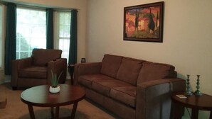 A living room with a brown sofa, a brown armchair, and a round coffee table. There is a painting on the wall and a window with curtains. - The Southwest Place 2203 (Ozark)