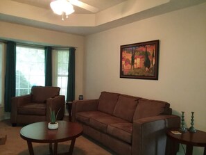 A living room with a brown sofa, a brown armchair, and a round coffee table. There is a painting on the wall and a window with curtains. - The Southwest Place 2203 (Ozark)