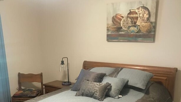 A bedroom with a wooden bed, a nightstand, a chair, a lamp, and a painting on the wall. - The Southwest Place 2203 (Ozark)
