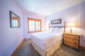 A bedroom with a large bed, two bedside tables with lamps, a dresser, a window with blinds, and a framed picture on the wall.
