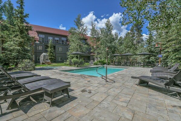A pool area with lounge chairs and umbrellas, surrounded by trees and a building with a red roof.