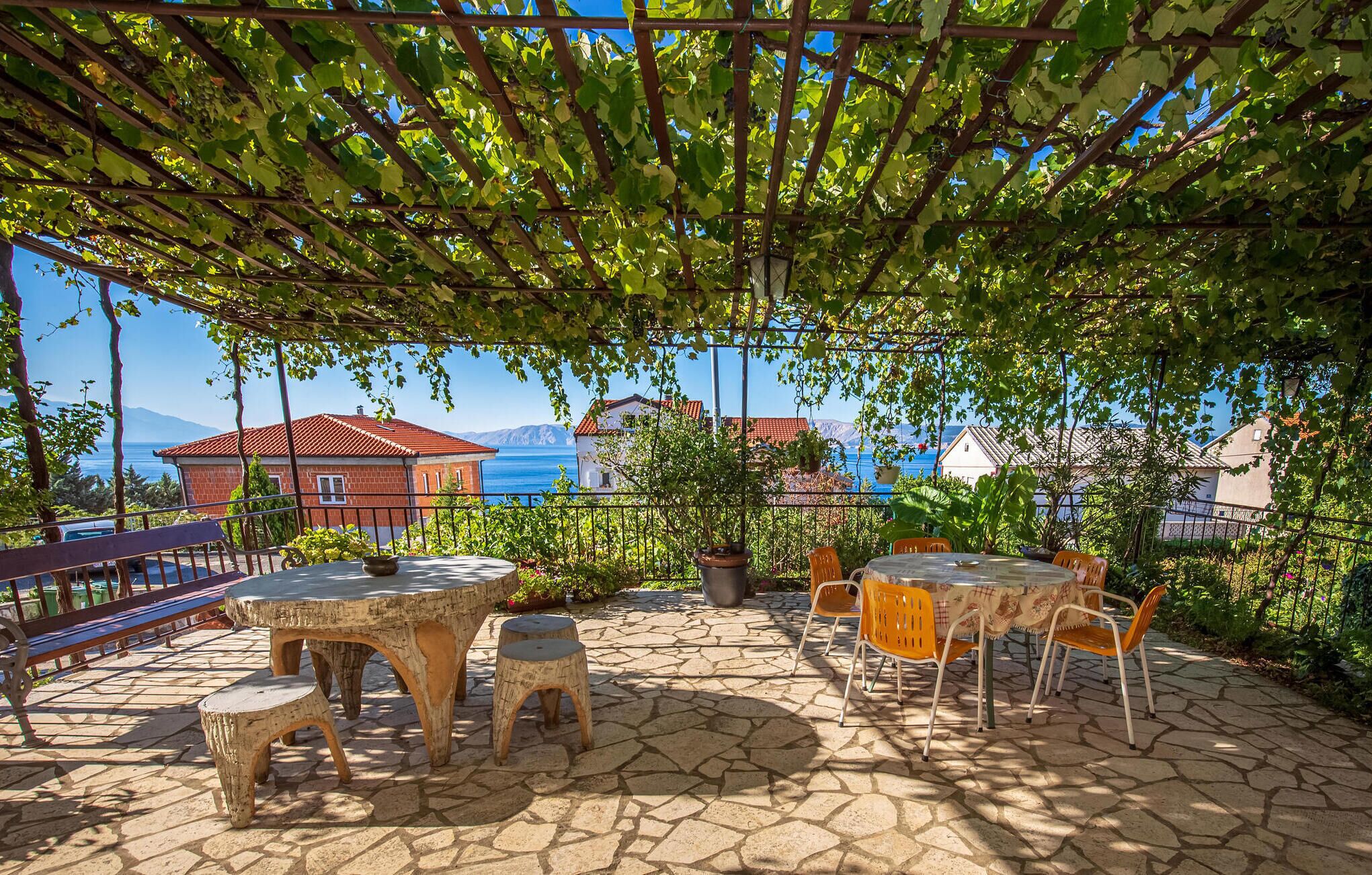 Un patio al aire libre con piso de piedra, mesas y sillas de madera, y una pérgola cubierta de hiedra.