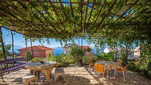 Une terrasse extérieure avec un sol en pierre, des tables et des chaises en bois, et une pergola couverte de plantes grimpantes.