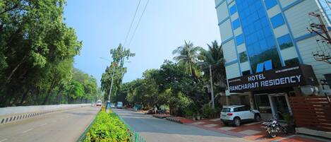 A hotel named Ramcharan Residency with a modern building, a road with vehicles, and lush greenery.