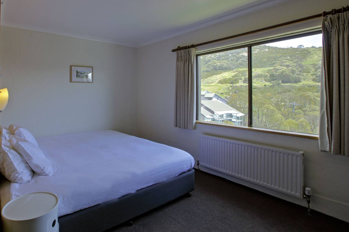 A bedroom with a large bed, a window with curtains, a radiator, and a picture on the wall.