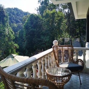 Un balcón con sillas de mimbre y una mesa de vidrio, con vista a una ladera boscosa.