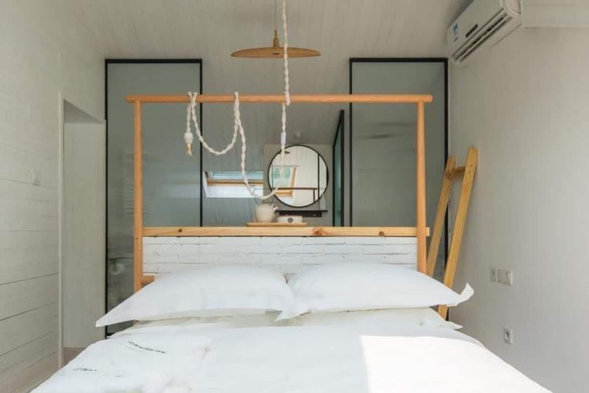 Un dormitorio con cabecera de cama de madera, ropa de cama blanca, una lámpara colgante, un espejo y una ventana con persianas.