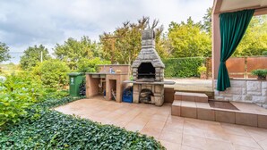 En udendørs terrasse med en stenbygget pizzaovn, et lille bord med en vanddispenser og en skraldespand.