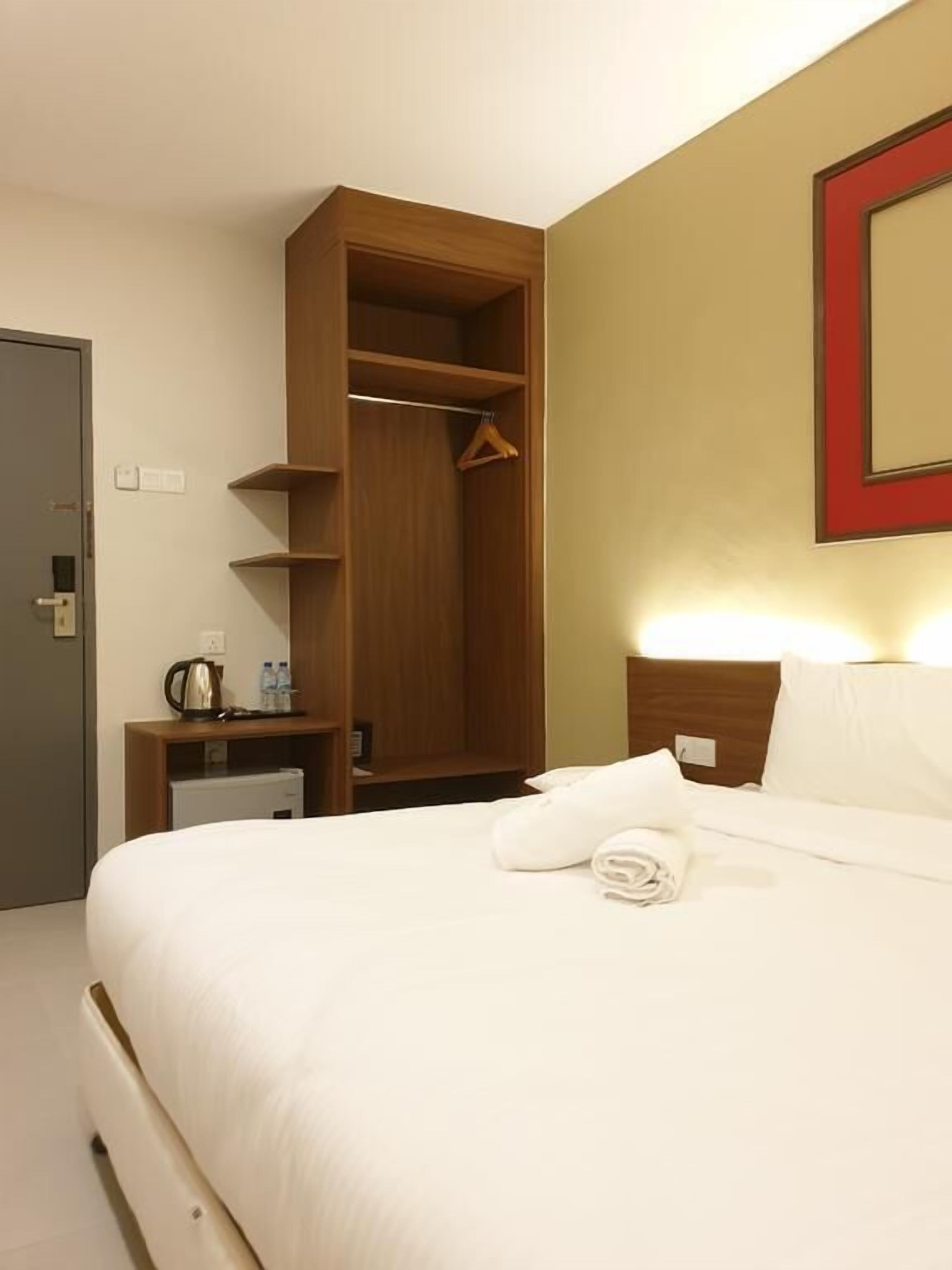 A hotel room with a bed, a wooden wardrobe, a bedside table with a kettle and water bottles, and a red-framed mirror on the wall.