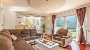 Ein modernes Wohnzimmer mit einem Ecksofa, einem weißen Couchtisch und einem gemütlichen Sessel. Im Hintergrund ist eine Küche erkennbar, und das Zimmer hat große Fenster mit Vorhängen.
