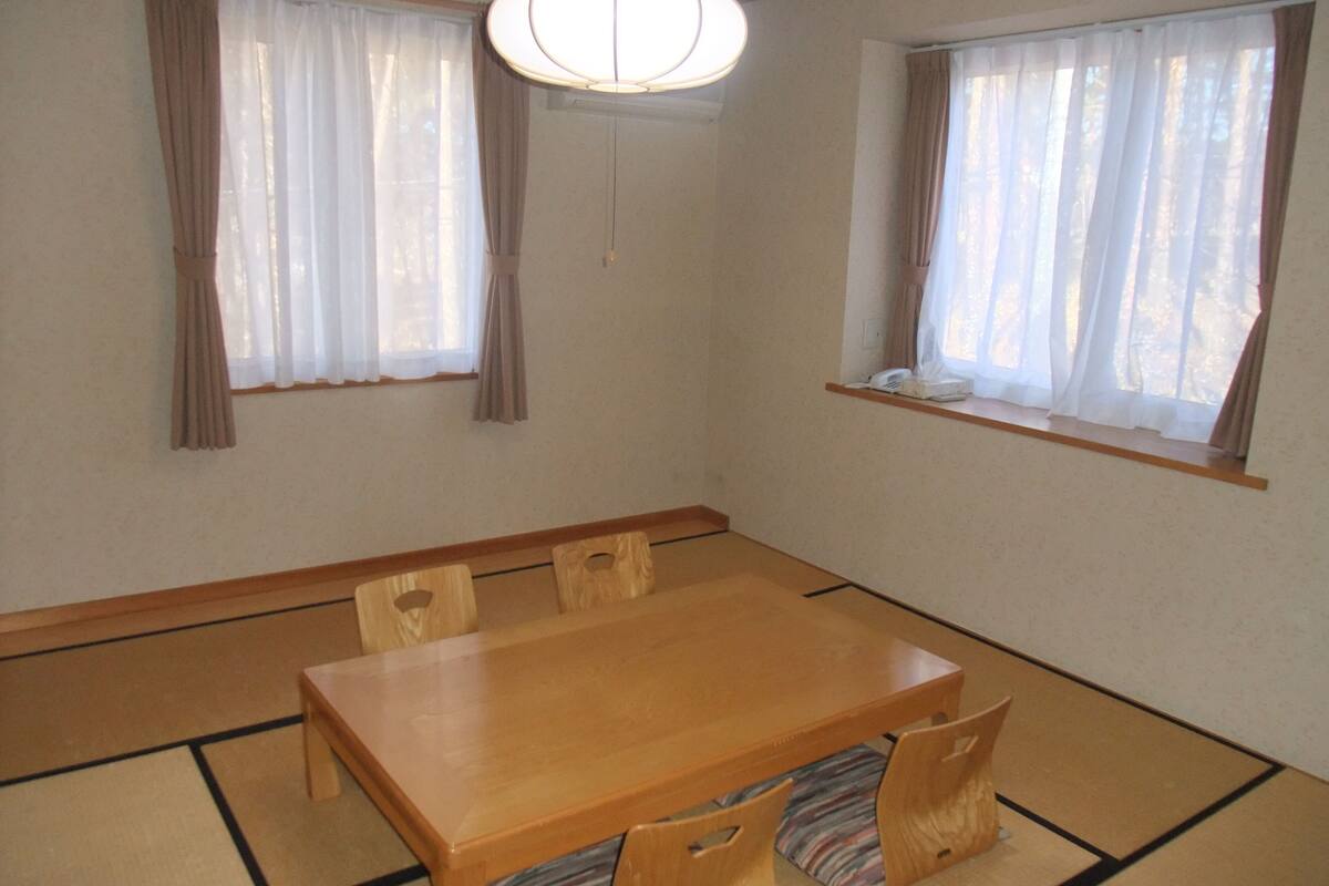 A traditional Japanese tatami room with a wooden table and chairs, two windows with curtains, and a hanging light fixture.