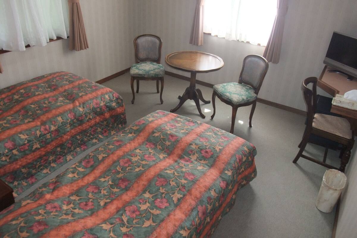 A hotel room with two single beds, a small round table, two chairs, a desk with a computer, and a TV.