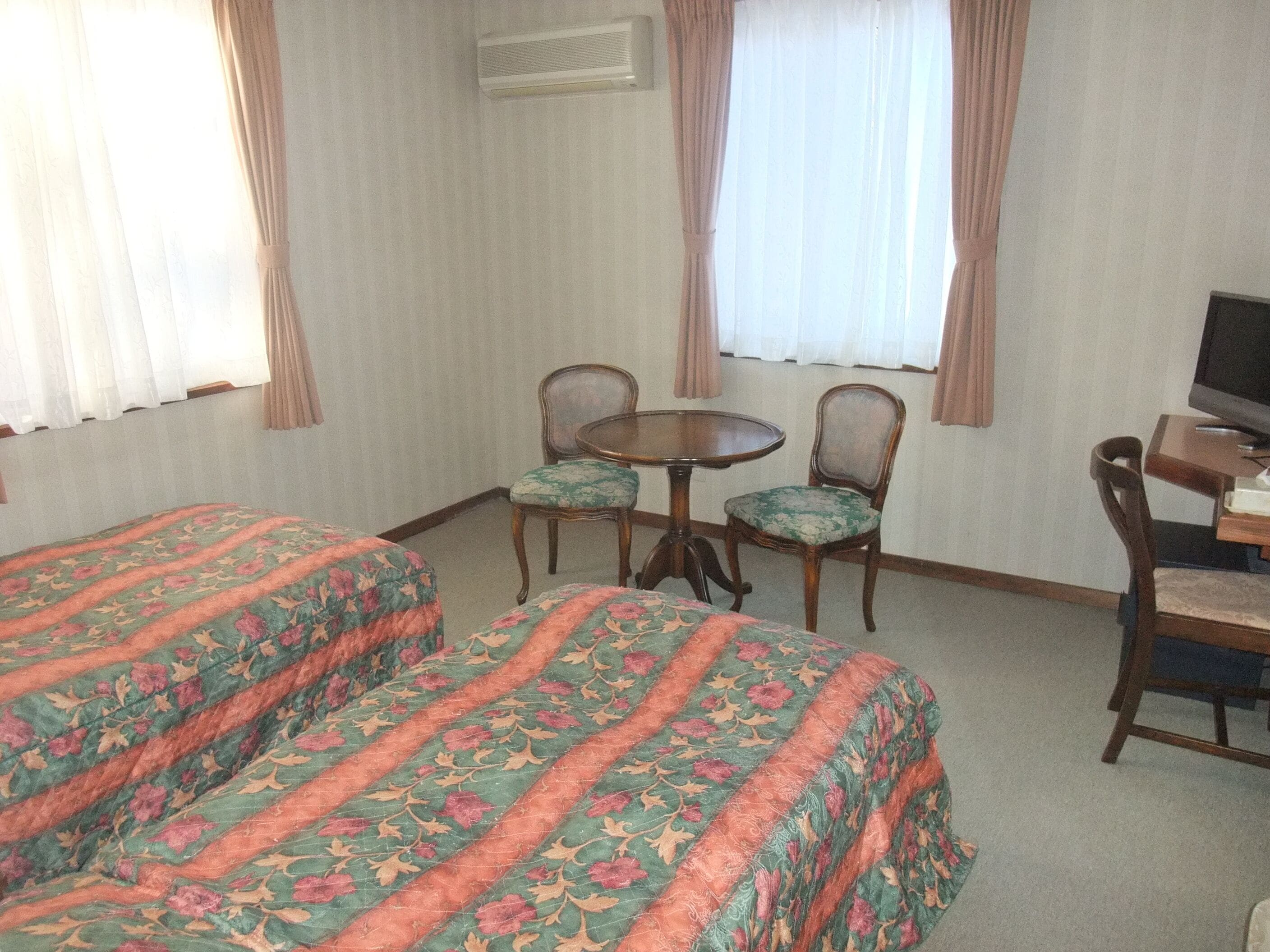 Hotellihuone, jossa on kaksi erillistä sänkyä, pieni pyöreä pöytä ja neljä tuolia, televisio puisen jalustan päällä sekä ikkuna, jossa on verhot.