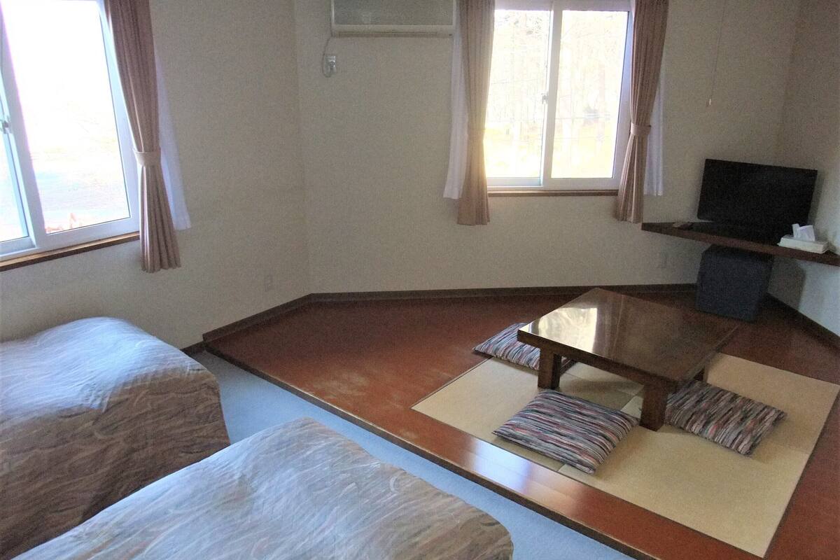 A room with tatami flooring, a low table with cushions, a television on a stand, and a bed with a white cover.