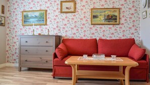 A room with a red sofa, wooden table, and floral wallpaper. There are two framed paintings on the wall.