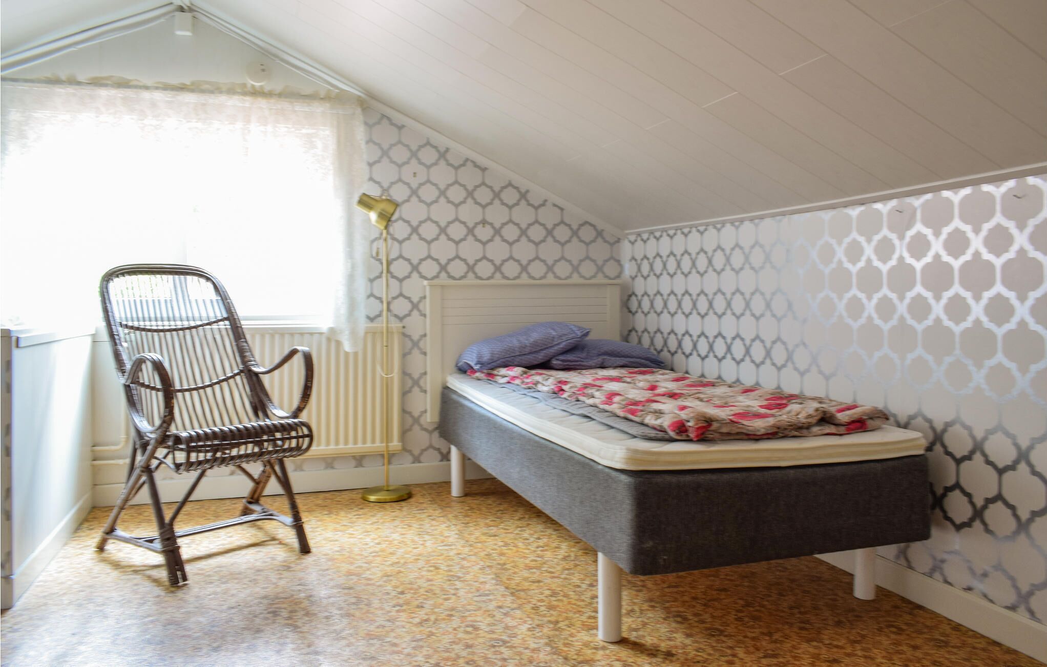 Ein kleines Zimmer mit einem Einzelbett, schrägen Wänden, gemustertem Wandpapier, einem Heizkörper, einem Stuhl und einer Nachttischlampe.