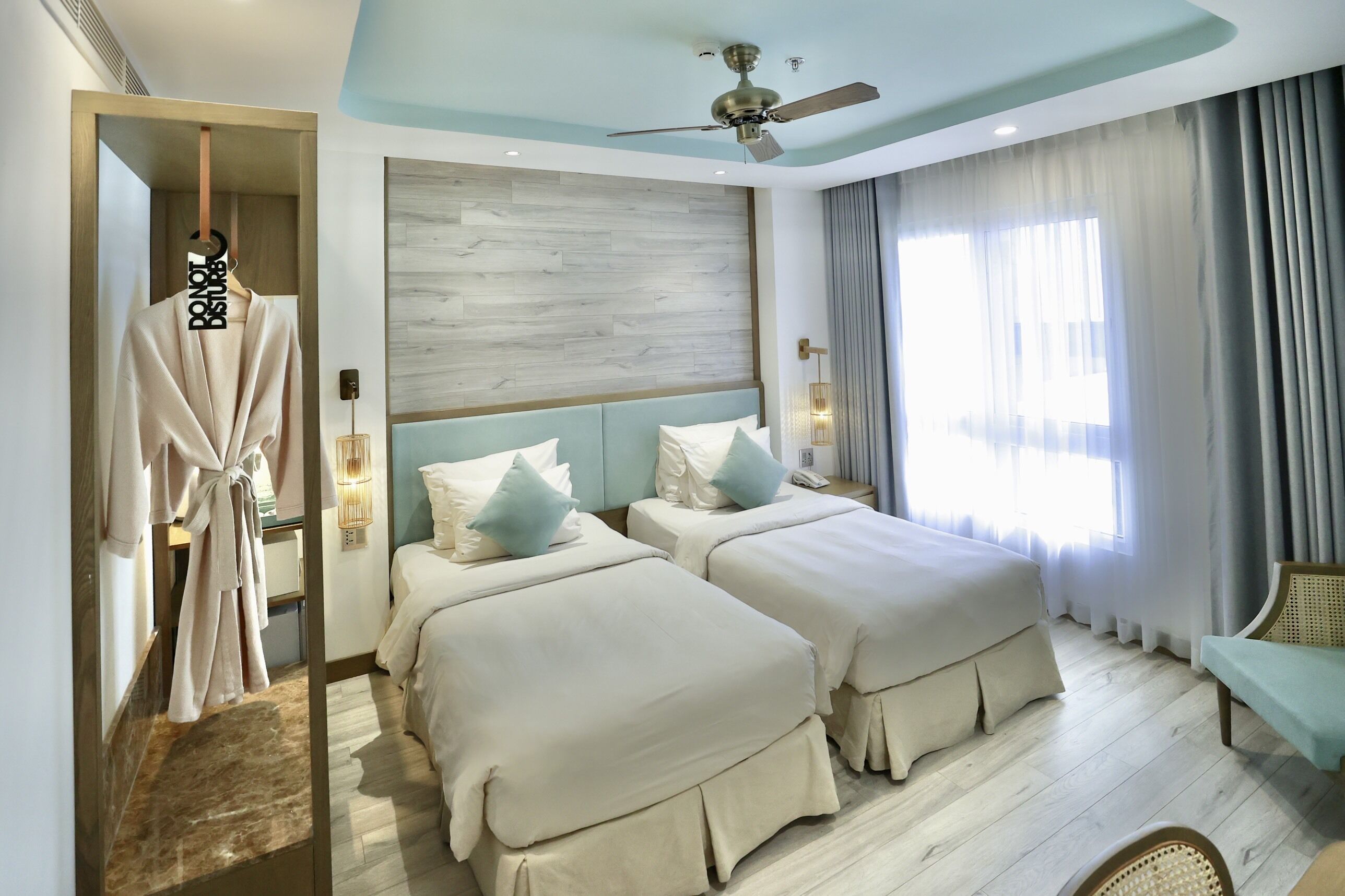 Une chambre d’hôtel avec deux lits, un porte-peignoir, un ventilateur de plafond et une vue sur l’extérieur par la fenêtre.