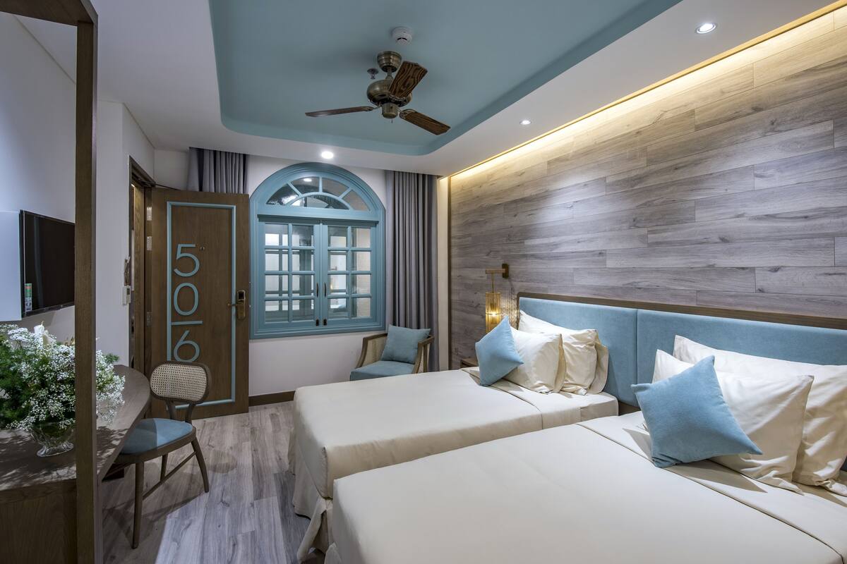 A hotel room with two beds, a ceiling fan, a wooden accent wall, and a window with curtains.