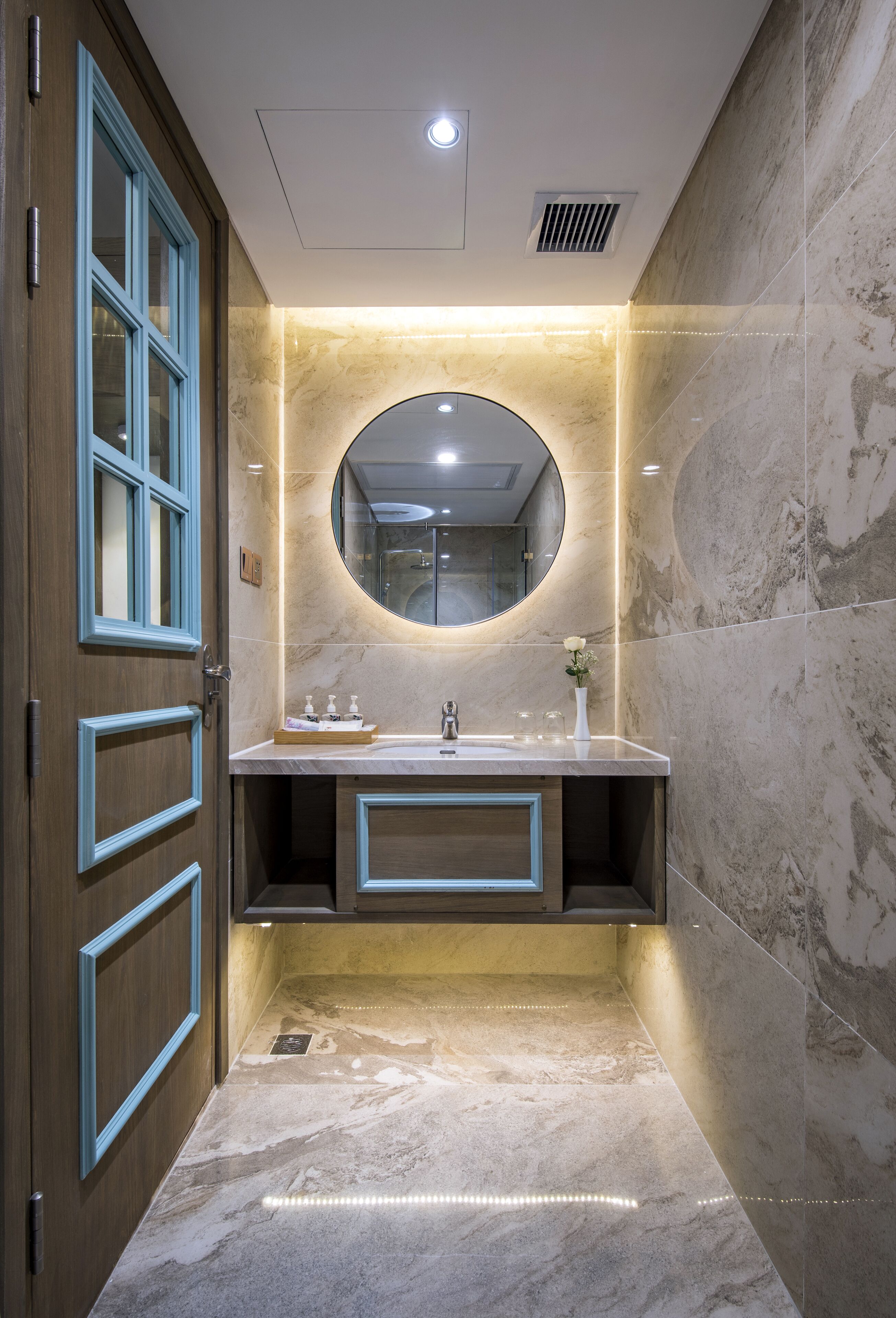 A bathroom with a marble floor, a wooden door with blue trim, a round mirror, and a vanity with a sink and toiletries.