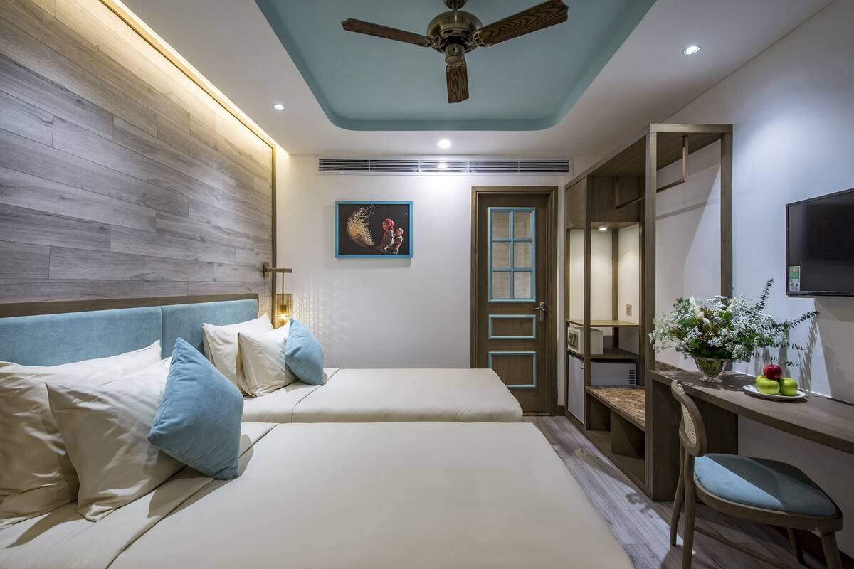 A hotel room with two beds, a desk, a chair, a ceiling fan, and a TV.