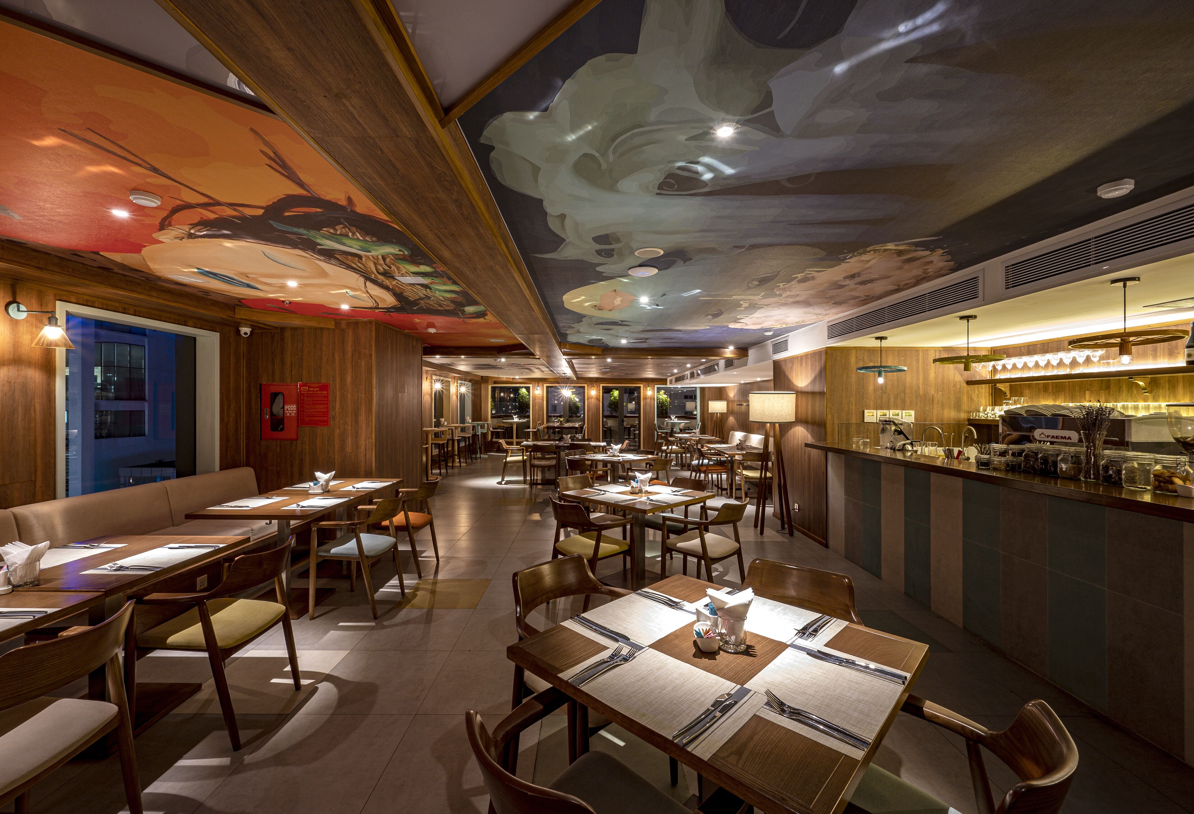 A spacious restaurant with multiple dining tables and booths, a bar area with a counter, and a ceiling mural.
