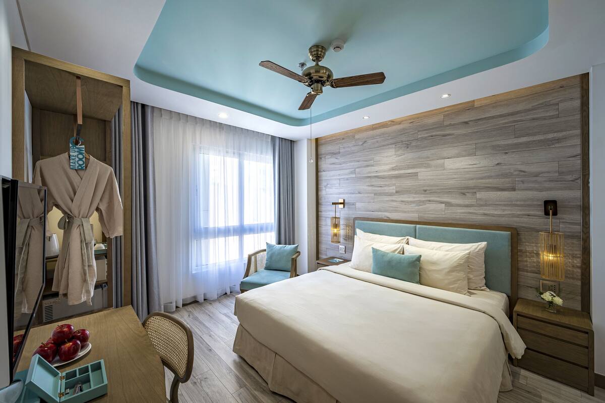 A hotel room with a large bed, a ceiling fan, a wooden headboard, a nightstand with a lamp, a chair, a robe hanging, and a window with curtains.