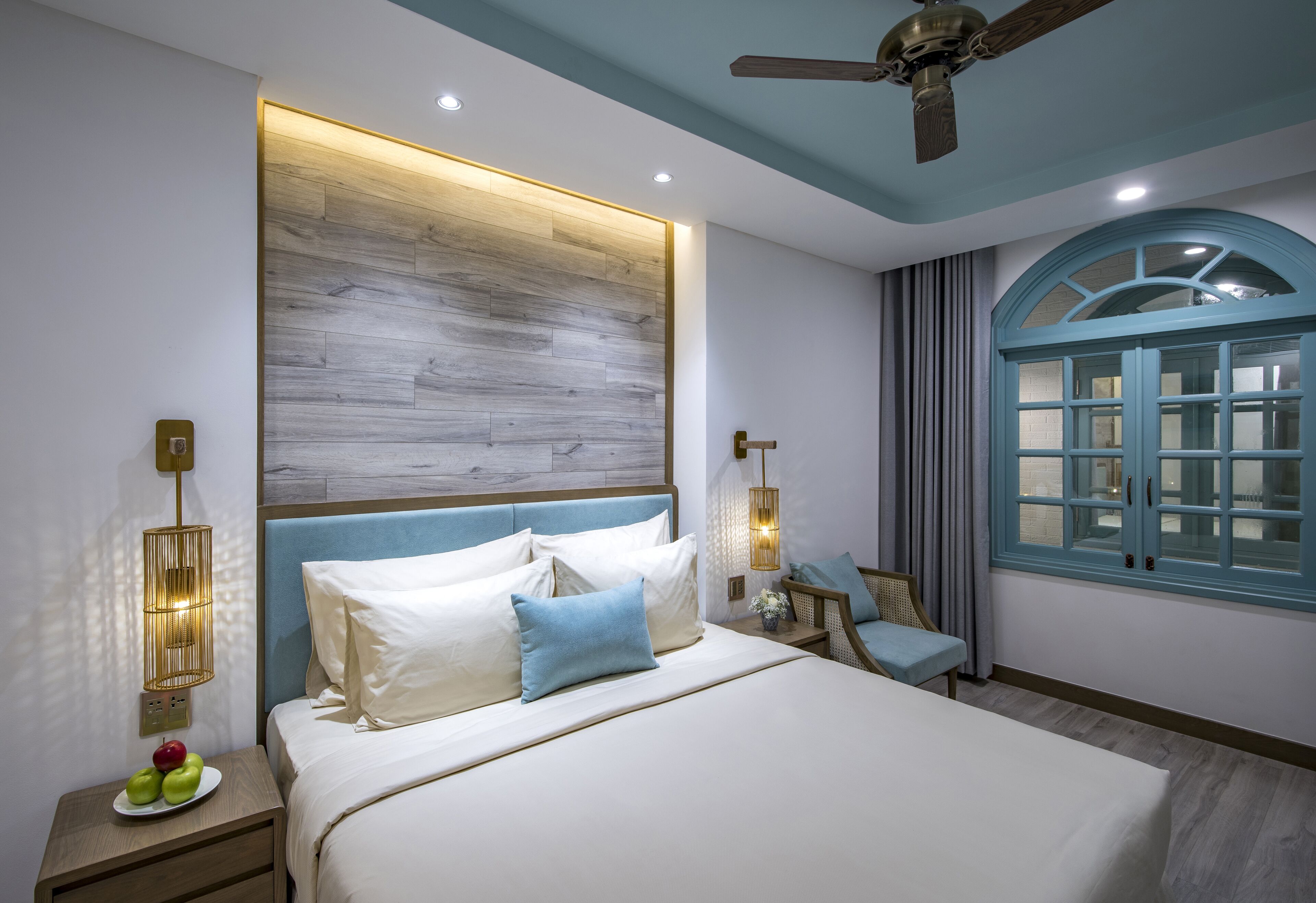 A modern bedroom with a large bed, a ceiling fan, a nightstand with a fruit plate, and a window with curtains.