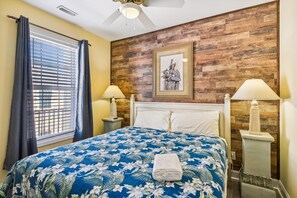 A hotel room with a bed, two bedside lamps, a ceiling fan, a window with blinds, and a wooden wall panel.