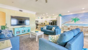A living room with a television, blue sofas, a coffee table, and a kitchen in the background. - Atlantis Villas 202 by Elliott Beach Rentals (NORTH MYRTLE BEACH)