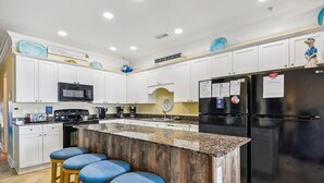 A modern kitchen with white cabinets, a black refrigerator, a microwave, and a coffee maker. There are three blue bar stools and a clock on the wall. - Atlantis Villas 202 by Elliott Beach Rentals (NORTH MYRTLE BEACH)
