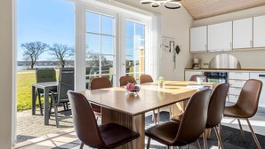 Comedor con una mesa grande y sillas, vista al exterior y una cocina con armarios blancos.