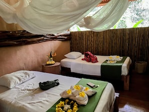 A room with two beds, each with white linens and a green bedspread. There is a small table with a vase of flowers and a red bag on one bed.