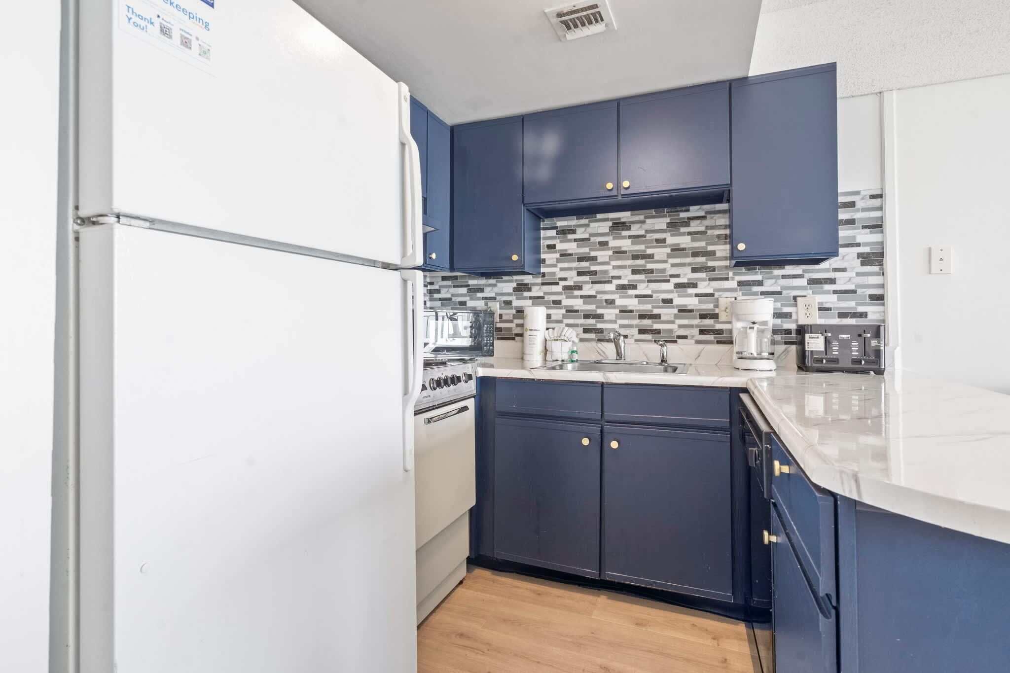 Een keuken met donkerblauwe kastjes, een witte koelkast, een betegelde spatwand en een aanrecht met diverse apparaten.