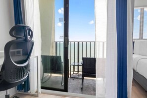 Une pièce dotée d’une porte-fenêtre coulissante donnant sur un balcon avec vue sur la mer, d’un fauteuil de bureau noir et d’un canapé blanc.