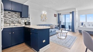 Una cocina moderna con armarios azul oscuro, una encimera blanca y una salpicadera. Se observa un rincón comedor con un sofá azul y una mesa redonda. Se ve un balcón con cortinas azules.
